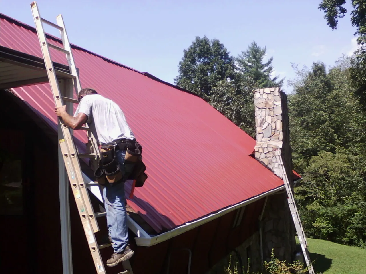 Roofer on a ladder beside a red metal roof and stone chimney — Soffit & Fascia Repair by Roofing Brothers in Richland